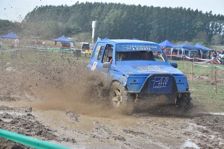 Suzuki Vitara azul avanzando en terreno fangoso, levantando barro, rodeado de carpas y vegetación.