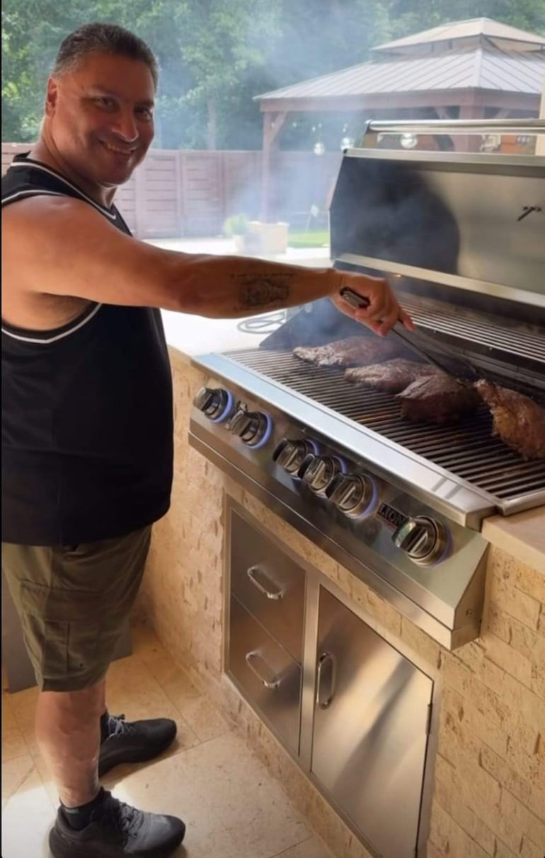 Hombre corpulento con camiseta sin mangas negra y pantalones cortos sonríe mientras asa carne en una parrilla, bajo un sol radiante.