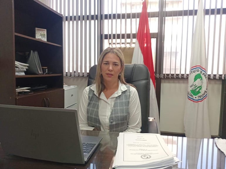 Cecilia Rodríguez con cabello lacio y camisa blanca, sentada en un escritorio con documentos y una computadora portátil, mira hacia la cámara.