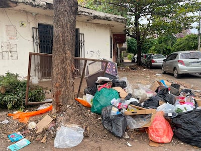 Montón de basura junto a un árbol en Asunción, con un marco de ventana inclinado y edificios de fondo.