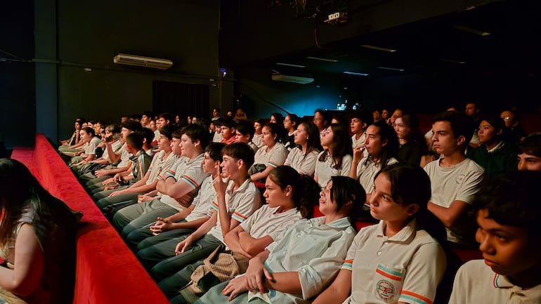 Grupo de jóvenes con camisetas blancas en un salón oscuro, escuchando atentamente la presentación.