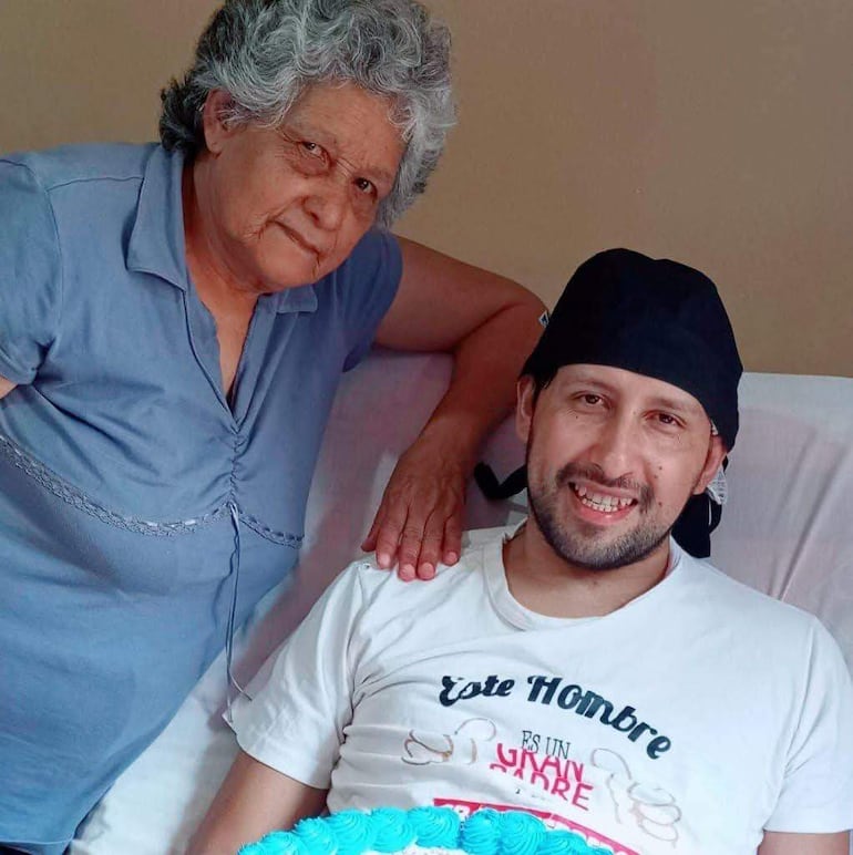 Hombre acostado en cama con camiseta que dice 'Este Hombre', acompañado por mujer de pie que lo apoya, ambiente acogedor.