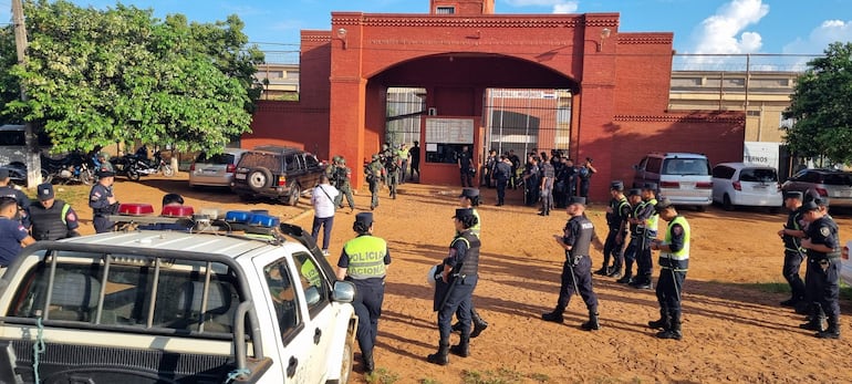 Funcionarios policiales uniformados conversan frente a un edificio de ladrillo rojo, junto a un vehículo policial con luces encendidas.
