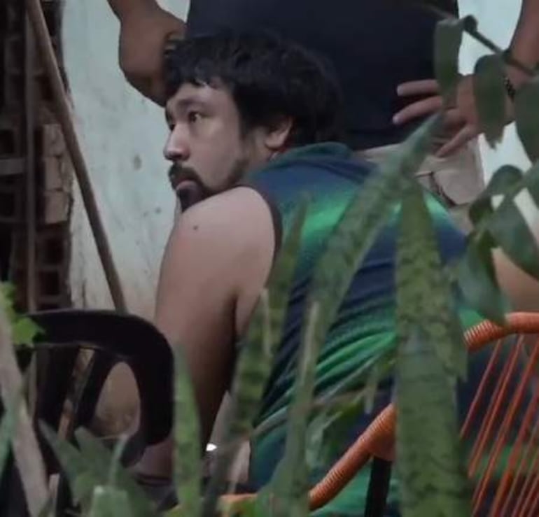 Hombre con cabello oscuro y rizado sentado en silla negra, rodeado de vegetación, en un entorno al aire libre.