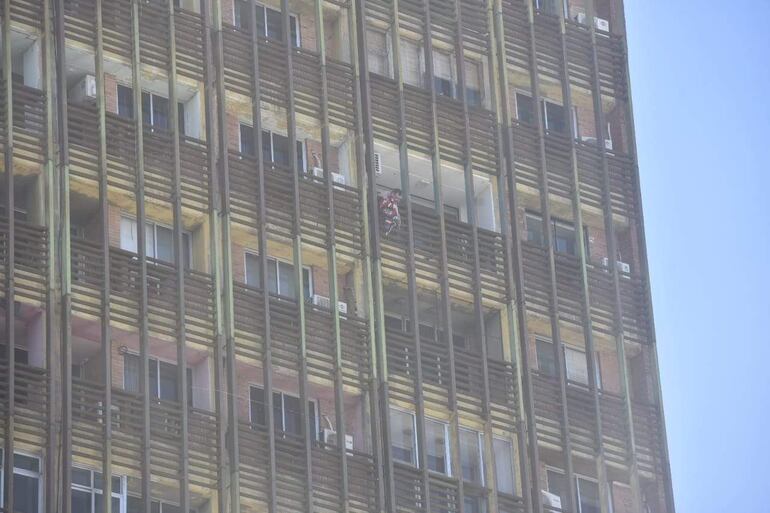 Persona en balcón vistiendo camiseta roja y blanca observa el edificio de Ahorros Paraguayos envuelto en humo.