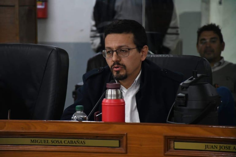 Miguel Sosa Cabañas con chaqueta y gafas habla, mientras Juan José escucha en mesas con botella de agua y termo rojo.