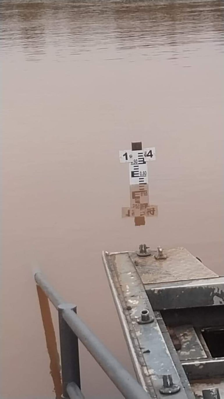 Marcador de nivel de agua parcialmente sumergido en agua marrón. Estructura metálica visible a la izquierda.