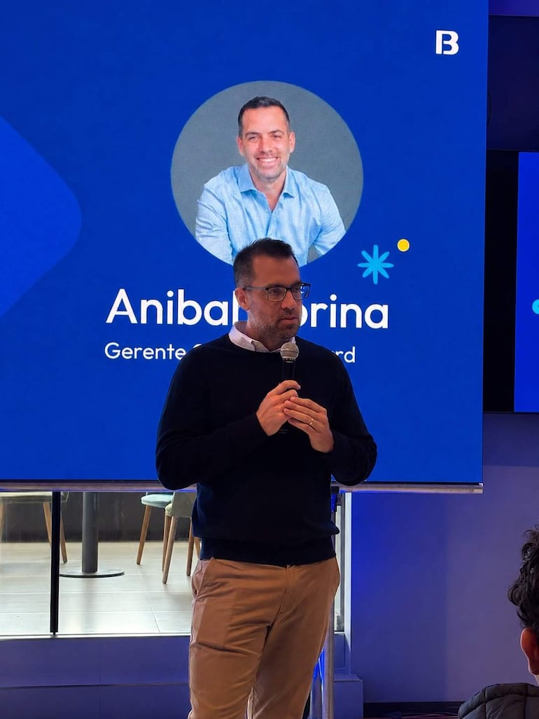 Aníbal Rina, con gafas y suéter oscuro, habla al micrófono frente a una audiencia en un evento corporativo.