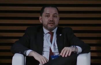 Luis Bello sentado en sillón blanco, vestido con traje oscuro y corbata roja, en un fondo de paneles de madera.