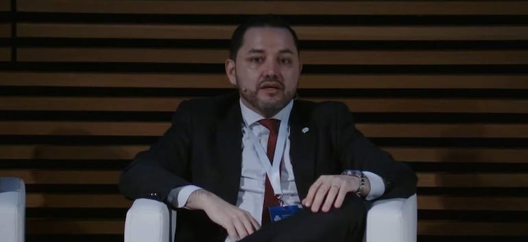 Luis Bello sentado en sillón blanco, vestido con traje oscuro y corbata roja, en un fondo de paneles de madera.