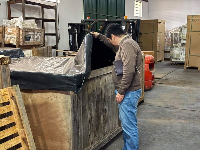 Hombre de chaqueta marrón y jeans claros inspecciona un contenedor en almacén con estanterías repletas de cajas.