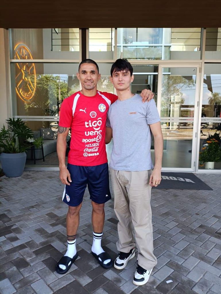 Víctor Gustavo Velázquez sonríe en camiseta roja de la selección paraguaya, junto a un hombre en camiseta gris, frente a un edificio moderno.