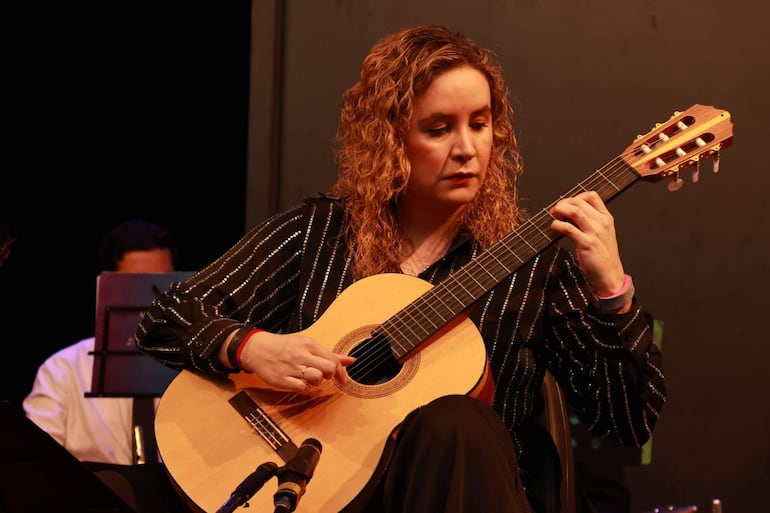 Daiana Ferreira Da Costa, con cabello rizado y camisa oscura, toca la guitarra mientras un hombre la acompaña en el escenario.