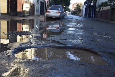 Calle urbana con charcos de agua y un automóvil plateado entre calles agrietadas y baches visibles.