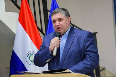 Alcides Riveros con saco azul y micrófono, hablando en un escenario con banderas de Paraguay y la Unión Europea.