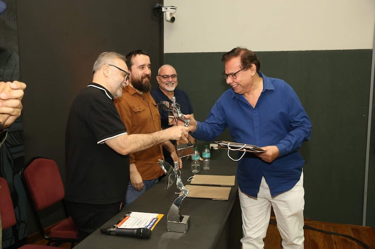 Hombres sonrientes en evento formal, uno levanta una mano en señal de alegría, sosteniendo un trofeo y documento.