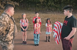 Seis niños con camisetas rojas y blancas, uno en el centro saluda, acompañado por un adulto en uniforme camuflado.