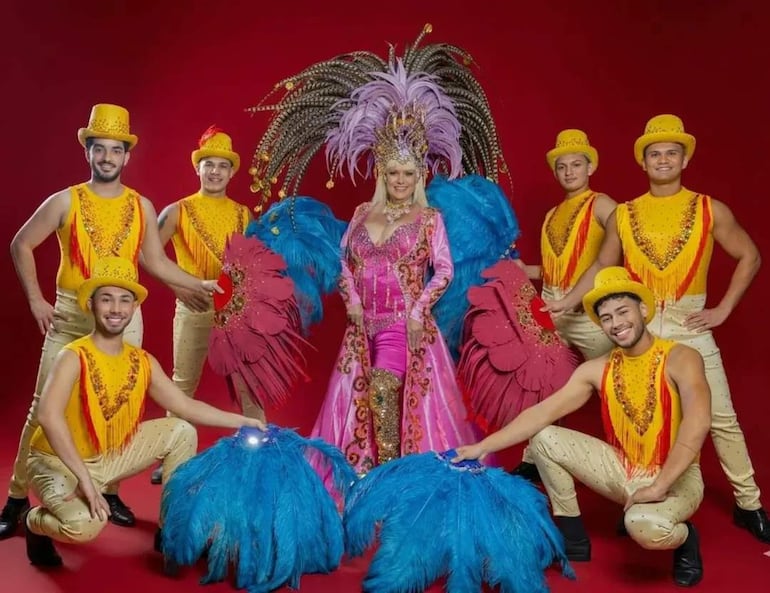 Bárbara Cibils en traje rosa y plumas azules, rodeada por seis hombres en camisetas amarillas y sombreros, en fondo rojo.