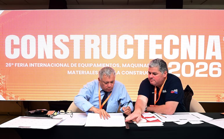 Dos hombres, uno en camisa clara y corbata, el otro en camiseta oscura, revisan documentos en una mesa frente a una pantalla.