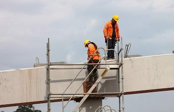 Dos trabajadores en el andamio, con cascos amarillos y chaquetas naranjas, trabajan en una estructura elevada de concreto bajo un cielo nublado.