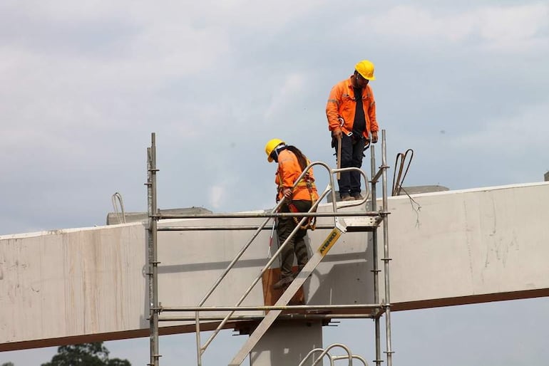 Dos trabajadores en el andamio, con cascos amarillos y chaquetas naranjas, trabajan en una estructura elevada de concreto bajo un cielo nublado.