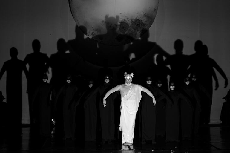Bailarín en toga blanca y coronita de hojas, rodeado por grupo en trajes negros, todos en escena teatral con fondo decorativo.
