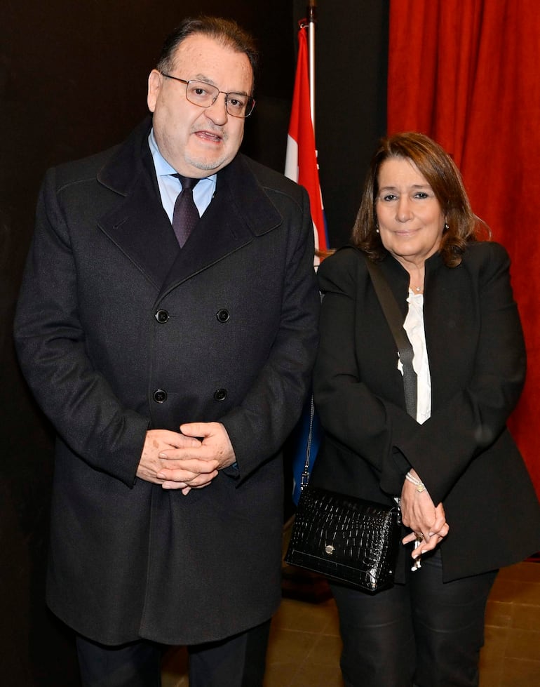 Hombre de abrigo negro conversa con mujer de conjunto negro en un entorno formal con fondo de cortina roja y bandera paraguaya.