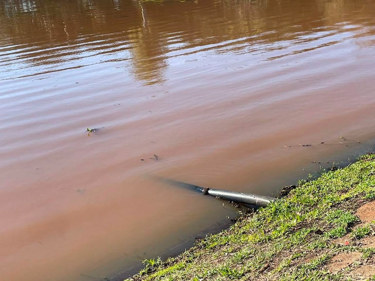 Cuerpo de agua marrón con una tubería sumergida y césped verde en la orilla, reflejando problemas de contaminación.