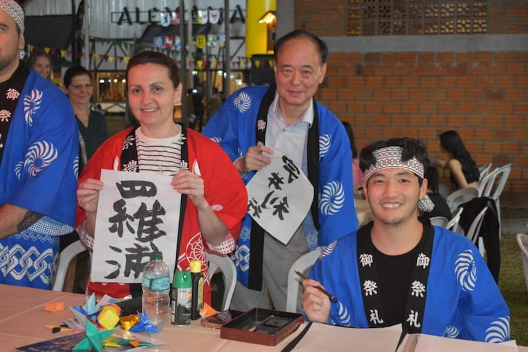 Cuatro jóvenes sonrientes, uno con camiseta azul y banda blanca, mujer en blusa de rayas y chaqueta roja, sosteniendo papeles en un ambiente festivo.