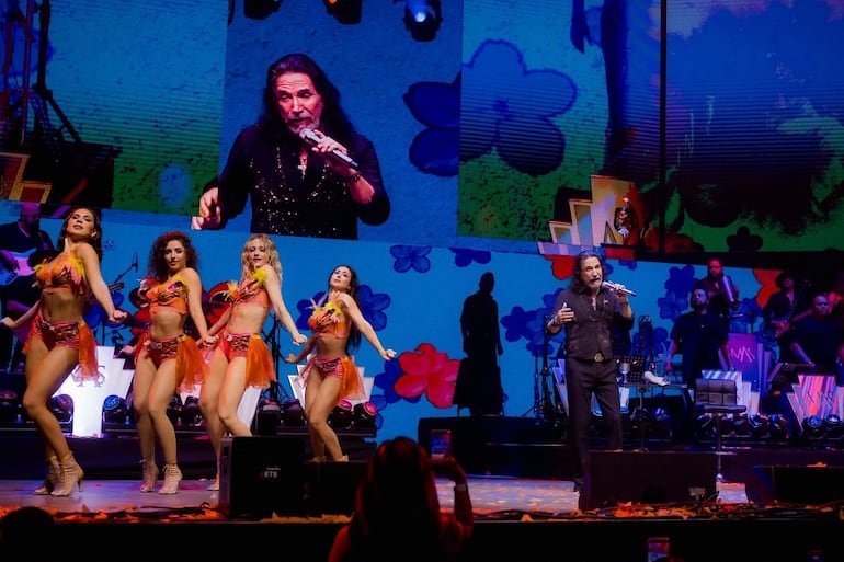 Marco Antonio Solís canta en escenario lleno de colores, acompañado de bailarinas con trajes de plumas y luces brillantes detrás.
