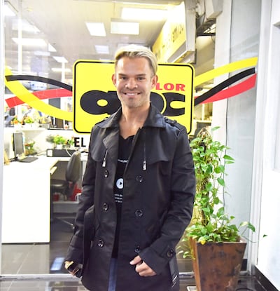 Persona de cabello rubio y abrigo negro sonríe frente al logo de ABC Color, en un ambiente interior con plantas.