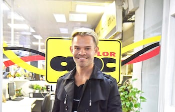 Persona de cabello rubio y abrigo negro sonríe frente al logo de ABC Color, en un ambiente interior con plantas.