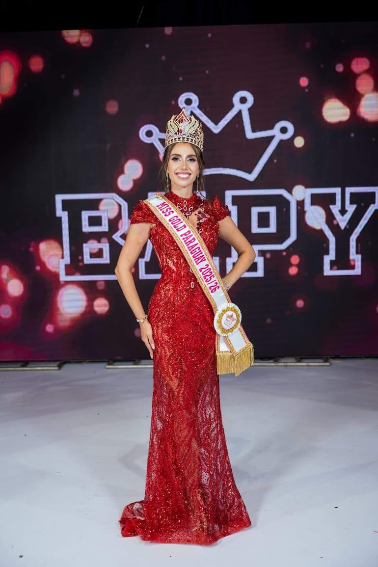 Natasha Reckziegel con vestido rojo y corona, sosteniendo cetro y banda de Miss Gold Paraguay en un escenario iluminado.