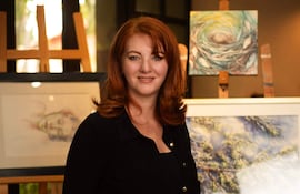 Monika Schuchardt sonríe con blusa negra, rodeada de obras de arte abstractas y paisajísticas en galería iluminada.