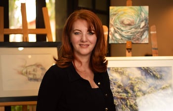 Monika Schuchardt sonríe con blusa negra, rodeada de obras de arte abstractas y paisajísticas en galería iluminada.