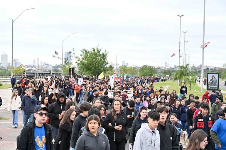 Grupo de jóvenes marchando con banderas y carteles, visten abrigos oscuros y camisetas coloridas, mostrando expresiones tranquilas y algunas tristes.