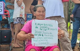 Mujer mayor con camiseta rosa sosteniendo cartel sobre transporte urbano, rodeada de manifestantes en espacio público.