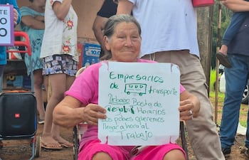 Mujer mayor con camiseta rosa sosteniendo cartel sobre transporte urbano, rodeada de manifestantes en espacio público.