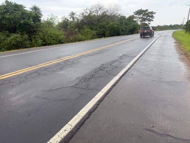 Carretera en mal estado con un vehículo oscuro en asfalto mojado, rodeada de vegetación densa.