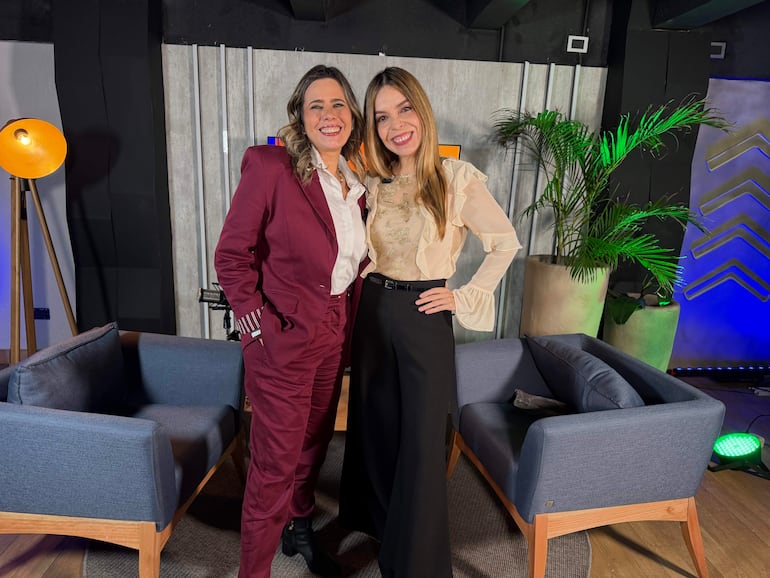 Kattya González con traje burdeos y Belén Bogado en blusa clara, felices en un set de grabación con sillones azules y plantas de fondo.