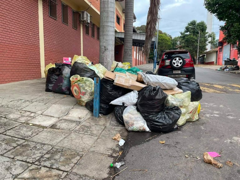 Bolsas de basura negras y amarillas acumuladas en la acera, con un automóvil rojo estacionado al lado.