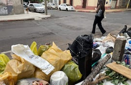 Persona con cabello largo y ropa oscura camina por la acera llena de basura, mientras un perro observa desde el otro lado.