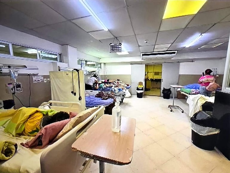 Ambiente hospitalario con camas ocupadas y personal organizando ropa, en una sala de cuidados.