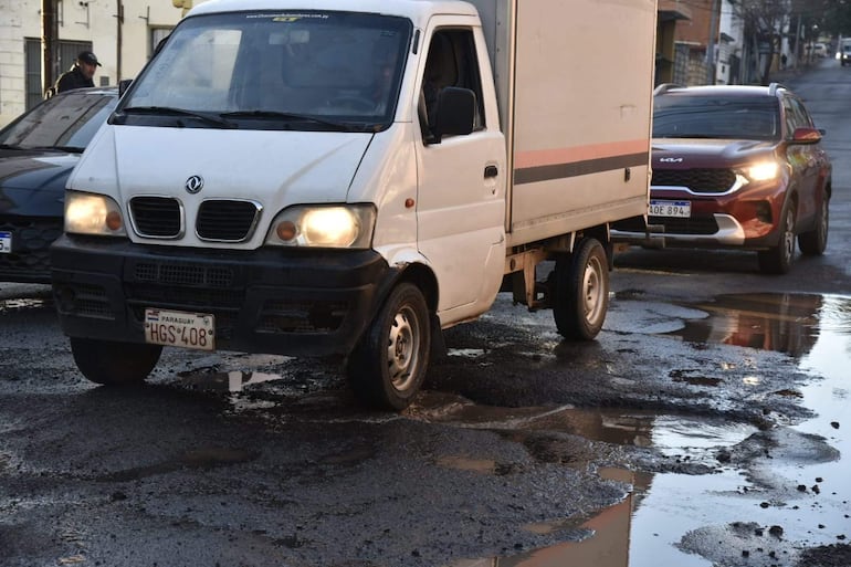 Camioneta blanca y auto rojo circulan en calle con baches y agua, sin personas visibles.