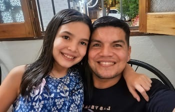 Hombre con camiseta negra y barba abraza a niña con blusa azul y estampado floral, ambos sonriendo en un interior luminoso.
