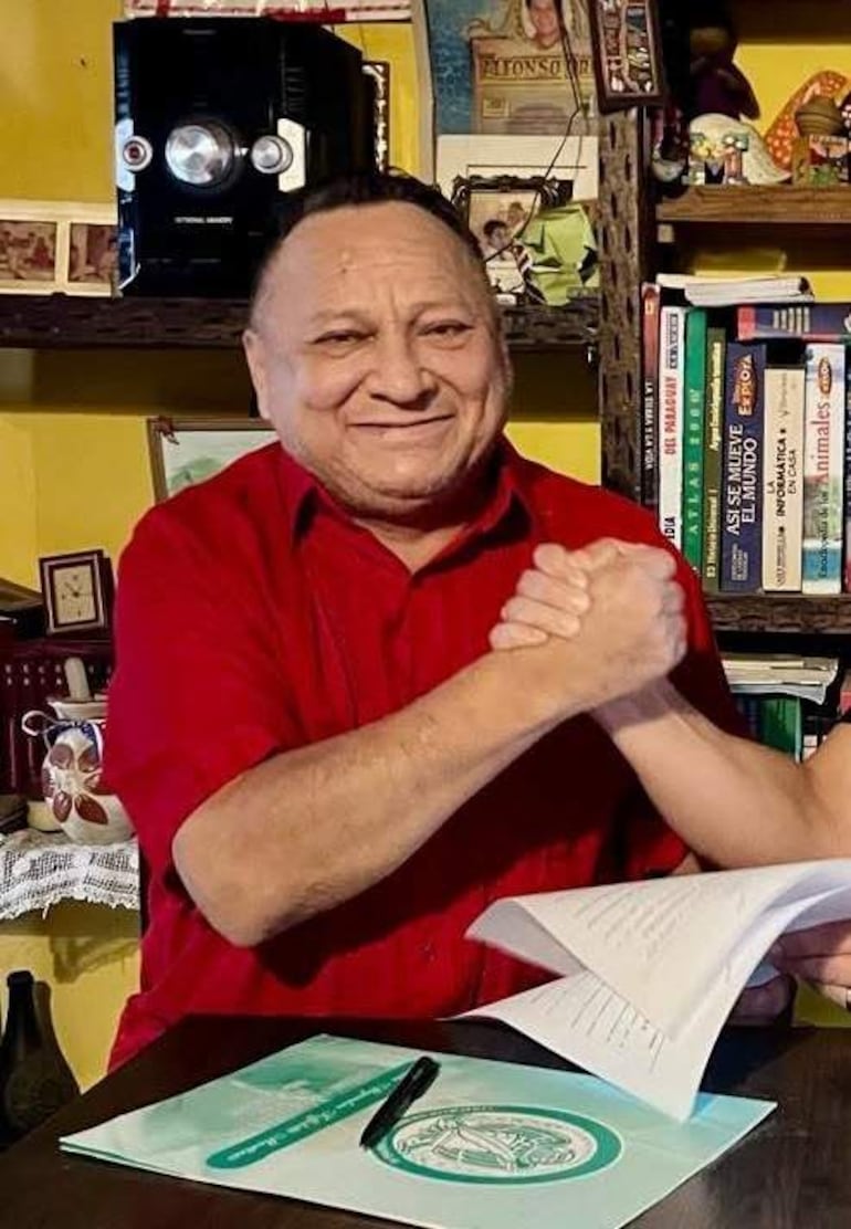 Alfonso Orella González, con camisa roja, sonríe y estrecha la mano en un ambiente acogedor, luciendo confianza y colaboración.