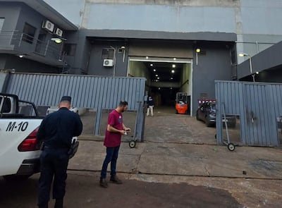 Policía de espaldas revisando vehículo M-10. Hombre en camiseta roja sosteniendo objeto, fondo de carga oscura y montacargas anaranjado.
