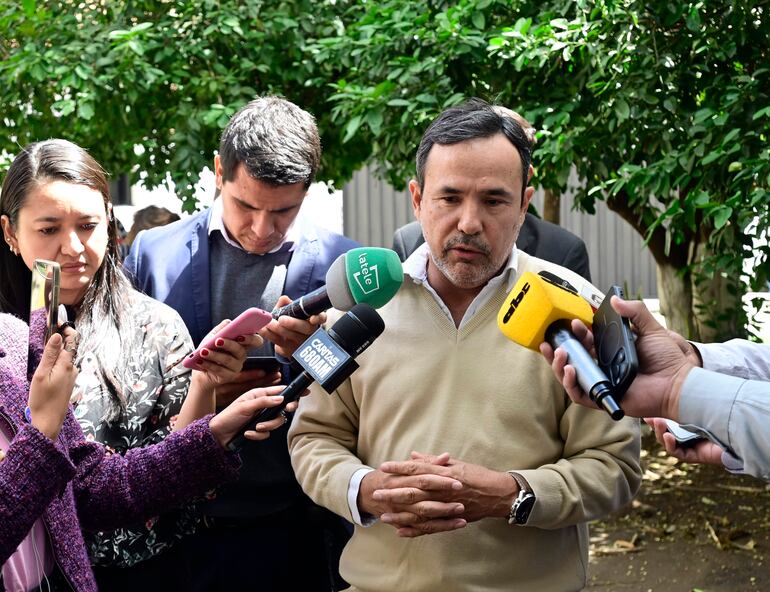 Emiliano Fernández en suéter claro, con expresión triste, rodeado de periodistas en un evento al aire libre con vegetación.