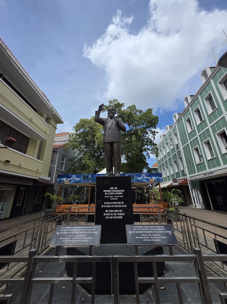 Escultura de Moisés Frumencio da Costa Gómez en gesto de saludo, rodeada de edificios coloridos y placas conmemorativas.