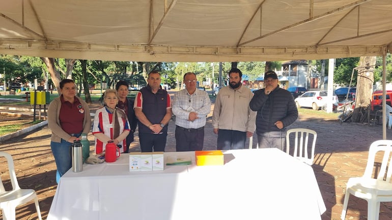 Grupo de siete personas escuchando a un hombre con micrófono, rodeados de objetos sobre una mesa en un ambiente al aire libre.